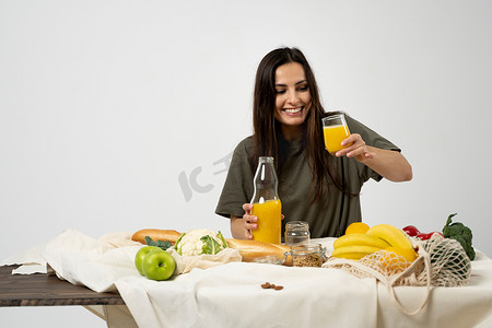蔬菜免费摄影照片_身穿绿色 T 恤的女人将玻璃瓶中的果汁倒在桌子上，桌子上放着网眼生态袋、健康的素食蔬菜、水果、面包、零食。