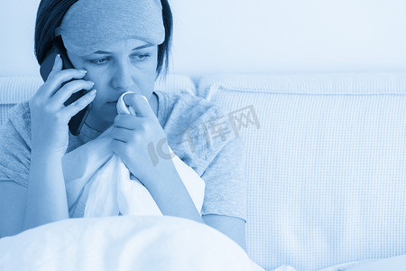 悲伤的女人戴着睡眠面具，坐在床上，用智能手机盖着羽绒被