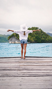 享受我们生活的这个美丽世界。后视照片显示一位面目全非的女性在假期中举起双臂站在木板路上俯瞰大海。