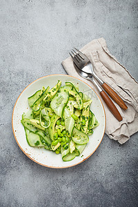 健康的素食绿色鳄梨沙拉碗，在陶瓷盘顶视图、灰石质朴的餐桌背景上配有切片黄瓜、毛豆、橄榄油和香草