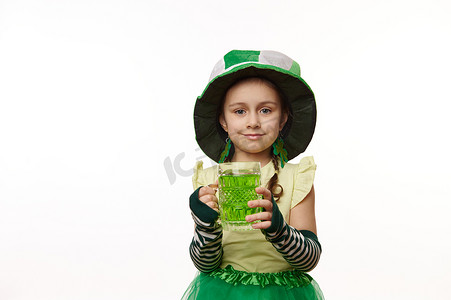 可爱的小女孩打扮成小妖精，戴着带三叶草叶子的帽子，在镜头前展示杯子里的绿色饮料。