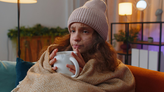 生病的少女戴着裹着格子花呢的帽子，坐在沙发上喝着热可可，冻得发抖