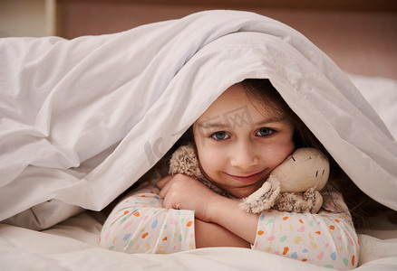 可爱的小女孩从柔软的毯子里挑出来，可爱地微笑着看着镜头，轻轻地拥抱她可爱的毛绒玩具