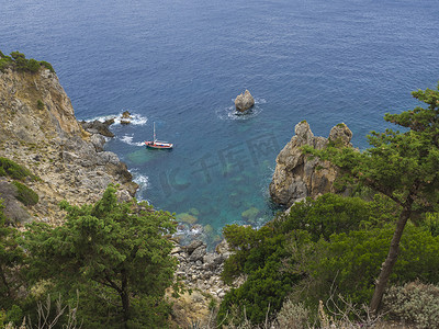 小山丘背景摄影照片_在 Paleokastritsa 湾的悬崖、树木和绿色山丘上欣赏小红船、夏季多云的天空、科孚岛、克基拉、希腊