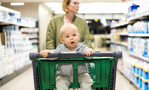 妈妈推孩子摄影照片_在超市杂货店的部门过道里，妈妈推着购物车带着她的婴儿男婴。