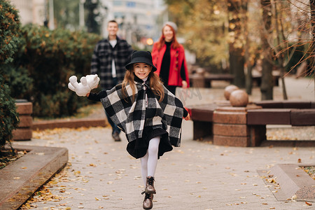 父母起跑摄影照片_一个小女孩和她的父母一起跑到秋天的城市。一个时尚的三口之家漫步在秋天的城市