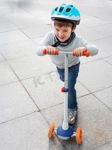 戴着头盔的蹒跚学步的人在停车场骑着滑板车，停着自行车。