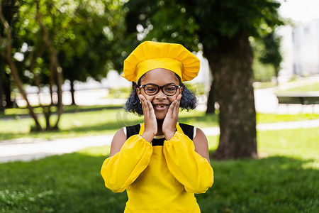 黑人厨师摄影照片_身穿厨师帽和黄色围裙制服的黑人非洲儿童厨师女孩震惊地双颊和惊喜。