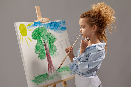迷人的女学生正在画架上用水彩笔绘画，站在灰色背景上。