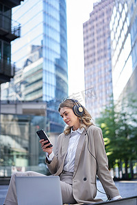 app新闻滚动摄影照片_穿着西装的企业女性，坐在城市里，用笔记本电脑听音乐，在手机上滚动新闻，午休时放松