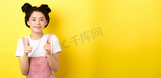 快乐的亚洲少女用手指指着镜头，微笑、鼓励或邀请你，赞美，赞美出色的工作，站在黄色背景上