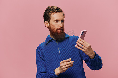 一位手持手机的红头发男子的肖像，他在手机上自拍和拍照，在粉红色背景下微笑，博主