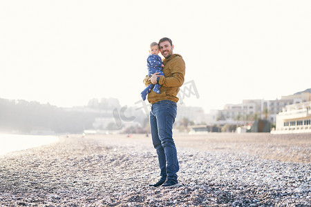 站在卵石滩上，身穿棕色夹克和牛仔裤的微笑爸爸抱着一个穿着蓝色工装裤的小孩