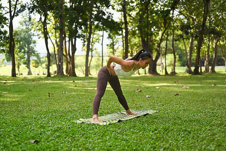 女健身瑜伽摄影照片_运动型瑜伽女练习瑜伽，站立潜水员瑜伽姿势，背景是绿叶树景观