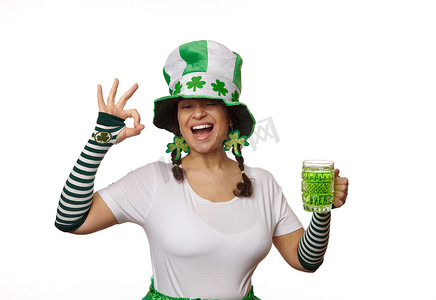戴着 Leprechaun 狂欢节帽子的漂亮女人，展示 Ok 的手势，在白色背景中摆出一杯爱尔兰绿色啤酒