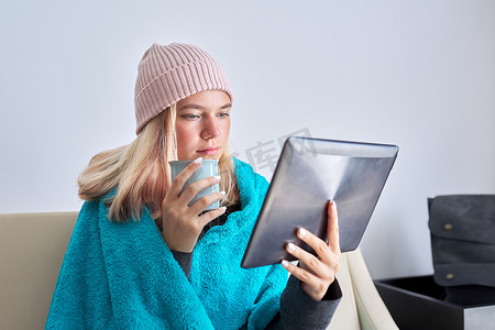 花纹茶杯摄影照片_戴着数字平板电脑茶杯的青少年学生坐在家里温暖的毯子下戴着帽子