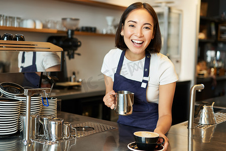 下单摄影照片_漂亮的年轻女咖啡师，做卡布奇诺，把拿铁艺术的蒸牛奶倒进杯子里，在柜台后面下单
