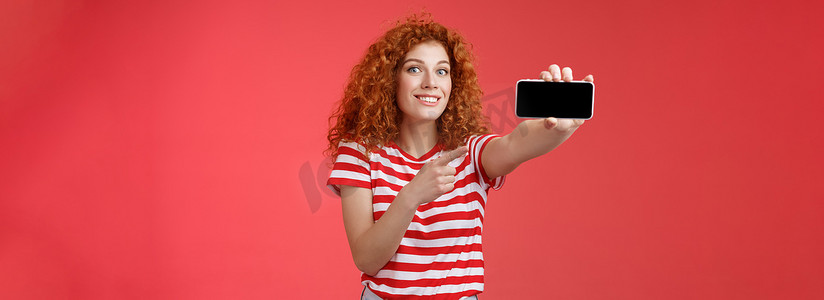 兴奋快乐好看的红发卷发女性展示水平智能手机屏幕指向显示小工具微笑高兴自豪击败朋友得分游戏站立红色背景