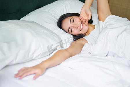 浪漫的亚洲女孩在舒适的卧室里醒来，躺在铺着白床单的床上，把手伸向空枕头
