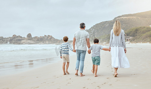 在一个轻松有趣的家庭暑假期间，快乐的白人父母和孩子们从后面牵着手沿着海滩散步。