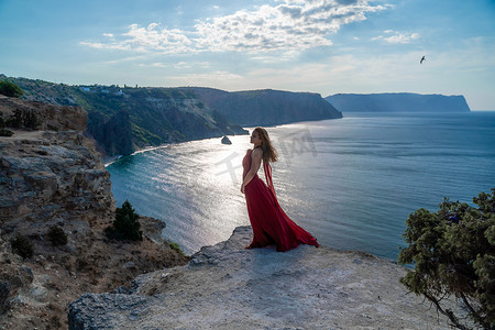 一位身穿红色长裙、头发飘逸的女孩站在海面的岩石上。