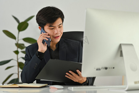 微笑的男性会计顾问拿着开放式办公室活页夹，坐在工作站时用手机聊天