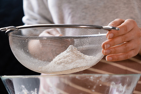 特写女人通过筛子将小麦粉筛入混合碗中，为黑色背景的蛋糕和面包店准备面团。