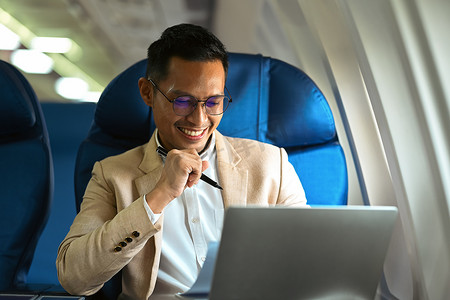 商务旅行期间身着优雅豪华西装、在飞机上使用笔记本电脑的微笑商人形象