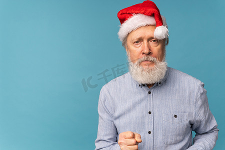 戴着圣诞帽的白胡子老人的画室肖像，手握拳头，用愤怒的表情复制空间看着相机 — 情绪和糟糕的圣诞情绪概念