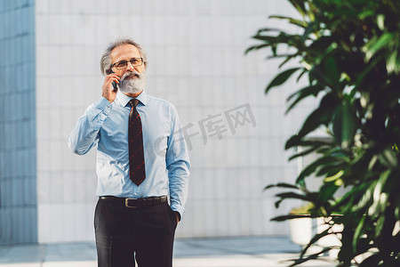 灰白的头发摄影照片_一位头发灰白、留着胡须的高级商人一边打电话一边走出办公室