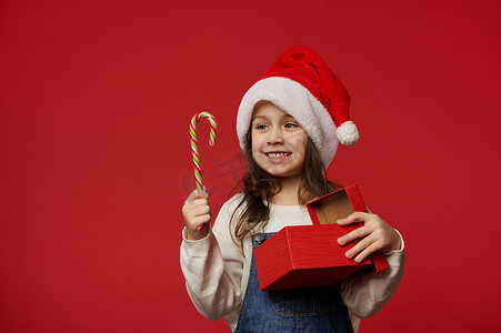 快乐可爱的小女孩戴着圣诞老人的帽子，在红色背景下拿着糖果手杖和圣诞礼物。