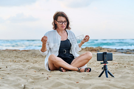 中年妇女坐在海滩上，用智能手机使用视频通话
