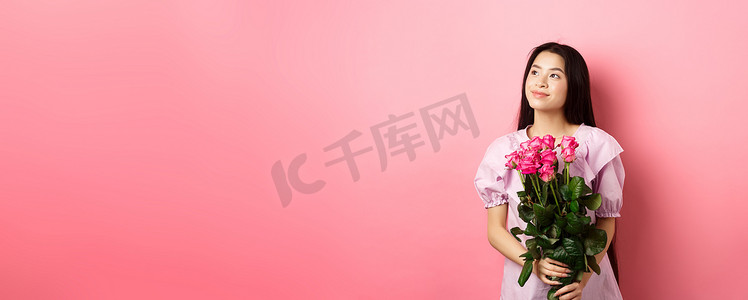 穿着可爱裙子的亚洲少女看着空荡荡的标志看起来很浪漫，拿着情人节鲜花礼物，从情人那里收到一束玫瑰，站在粉红色的背景上