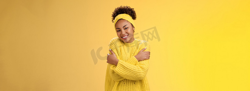 迷人的女性非洲裔美国女性可爱温柔的微笑，倾斜的头拥抱自己，感到舒适，爱轻轻地拥抱肩膀，穿着舒适的毛衣，开心地笑着黄色背景