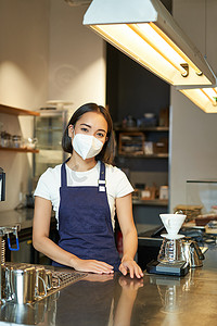 友善咖啡师的垂直照片，戴医用口罩的女职员，在柜台后与客户一起工作，在咖啡厅供应咖啡，穿着制服围裙