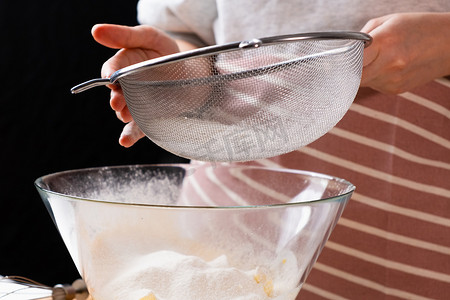 厨师面团摄影照片_特写女人通过筛子将小麦粉筛入混合碗中，为黑色背景的蛋糕和面包店准备面团。