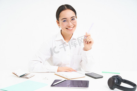 年轻的女售货员、亚洲女商人坐在办公室里，处理文件，桌上放着数字平板电脑和耳机，白色背景