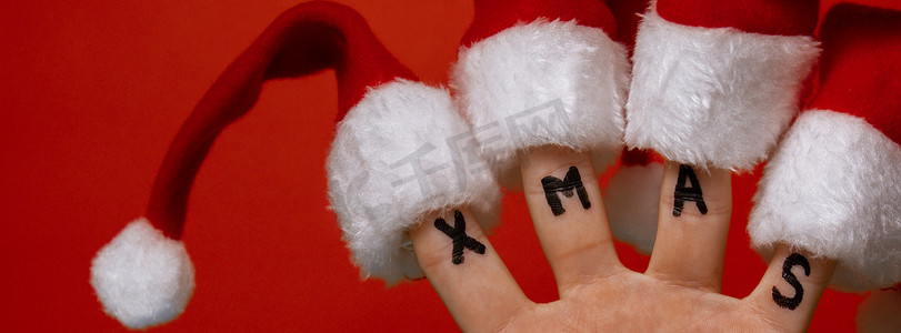 红色文字背景摄影照片_手指穿着圣诞老人红白帽子和红色背景上的文字 XMAS。