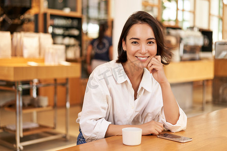 亚洲女商人的画像，坐在咖啡馆的桌子上，喝着咖啡，在智能手机应用上阅读，对着镜头微笑