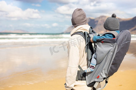 年轻的父亲在西班牙兰萨罗特岛法马拉多风的沙滩上背着他的婴儿男婴太阳在背包里。