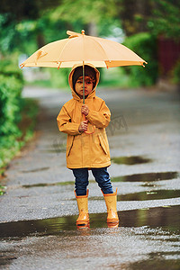 防水雨伞摄影照片_雨后穿着黄色防水斗篷、靴子和雨伞在户外玩耍的孩子