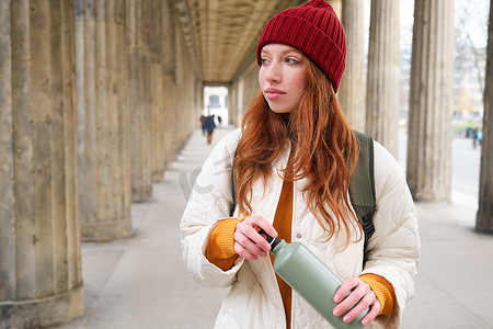 年轻的红发女孩手里拿着热水瓶，在城市里散步时给自己倒了一杯热饮。