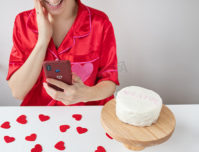 一个穿着红色衣服的快乐女孩拿着一个智能手机，放在一个樱桃盒里，上面有一颗心，并且很高兴收到来自亲人的短信、蛋糕和小红心放在桌子上。