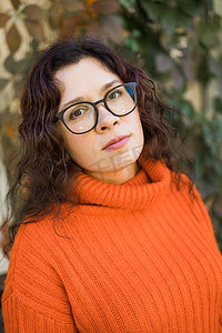 时尚橙色摄影照片_秋天的画像中，一位戴着时尚眼镜、身穿时尚橙色针织毛衣、背景为树叶的迷人年轻女子的画像。