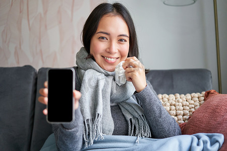 兴奋的年轻女性用手指指着智能手机，在手机上显示在线医生、医疗应用程序或 GP 联系方式，呆在家里生病、感冒