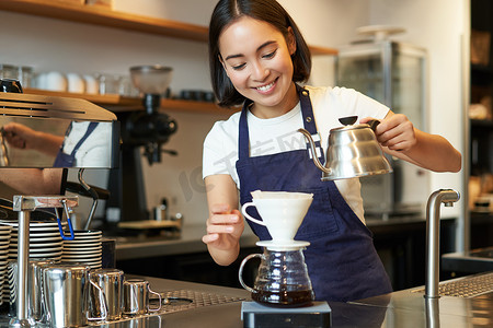 咖啡厅女孩摄影照片_可爱的黑发女孩咖啡师，咖啡厅工作人员从水壶里倒水，在柜台后面冲泡过滤咖啡，准备点餐