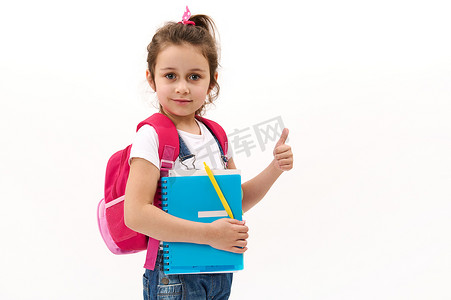 可爱的 5-6 岁小女孩，背着粉色背包，拿着课本，竖起大拇指看着相机。