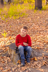 穿着红色连肩衣的快乐小家伙坐在秋天黄色公园的树桩上