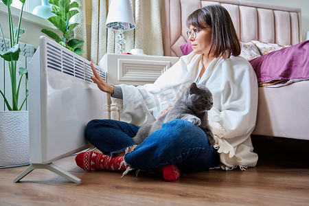 女人和猫坐在毯子下，在电加热器附近取暖