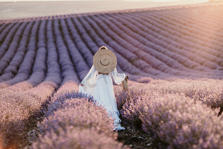 紫色氛围摄影照片_穿着白色连衣裙和草帽的快乐女人在日出时漫步穿过薰衣草田，沉浸在宁静的氛围中。